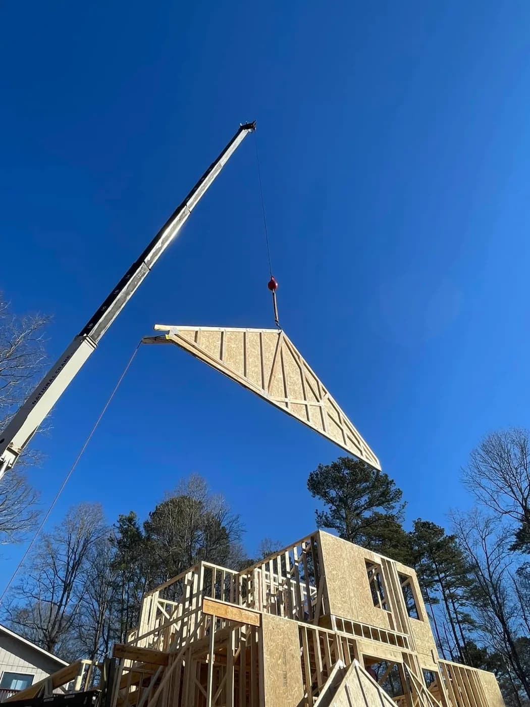 Crane lifting a roof truss over a house under framing against a clear blue sky